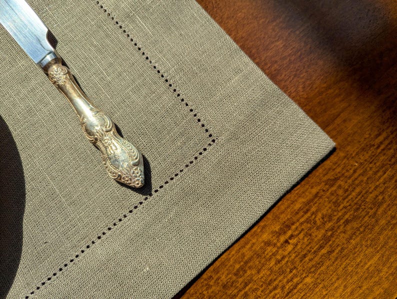 Puede incluir: Un cuchillo de mantequilla de plata con un mango adornado descansa sobre un mantel individual de lino beige. El mantel individual tiene un borde decorativo cosido.