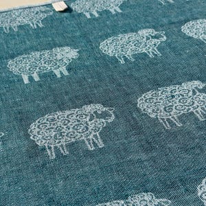 May include: A teal blue kitchen towel with a white repeating pattern of sheep. The sheep are stylised and have a curly wool design.