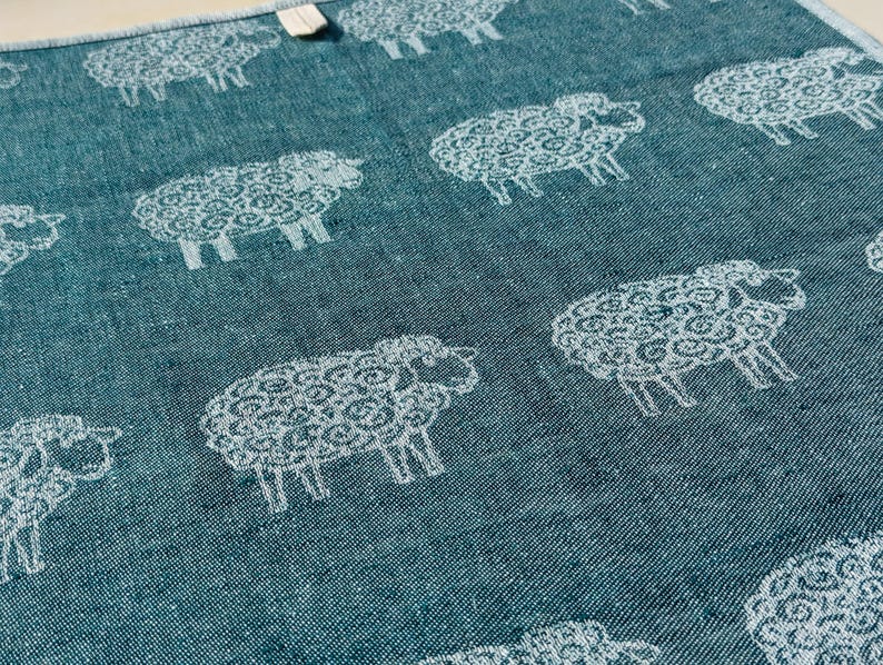 May include: A teal blue kitchen towel with a white repeating pattern of sheep. The sheep are stylised and have a curly wool design.