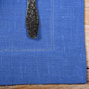 May include: A blue linen napkin with a decorative silver spoon on top. The napkin has a stitched border.
