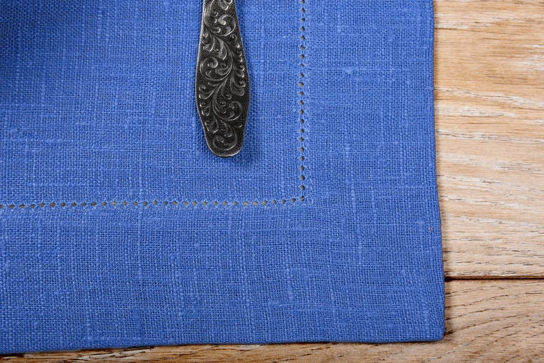 May include: A blue linen napkin with a decorative silver spoon on top. The napkin has a stitched border.