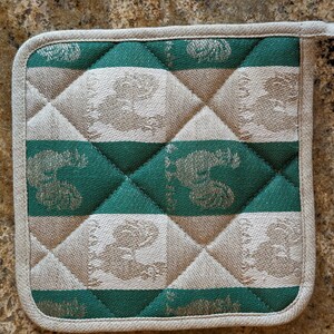 Puede incluir: Un agarrador de olla acolchado verde y beige con un patr&oacute;n repetido de p&aacute;jaros estilizados. El agarrador de olla tiene un borde blanco.