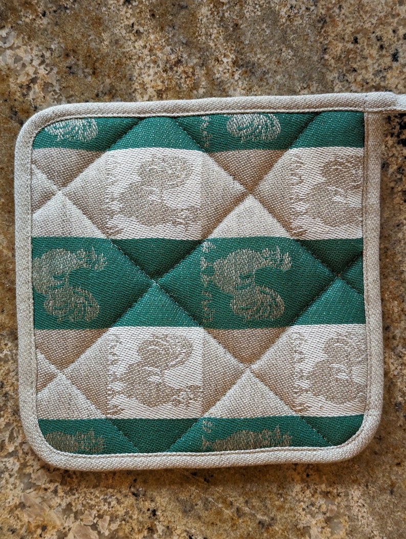 Puede incluir: Un agarrador de olla acolchado verde y beige con un patr&oacute;n repetido de p&aacute;jaros estilizados. El agarrador de olla tiene un borde blanco.