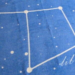 May include: A blue throw blanket with a white constellation design of Libra. The word "Libra" is written in white script in the bottom right corner.