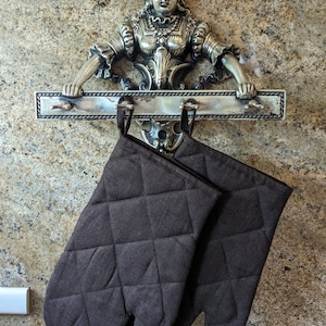 K&ouml;nnte beinhalten: Zwei braune, gesteppte Ofenhandschuhe, die an einem silbernen Metallgestell h&auml;ngen, auf dem sich eine dekorative Figur einer Frau in einem Kleid befindet.