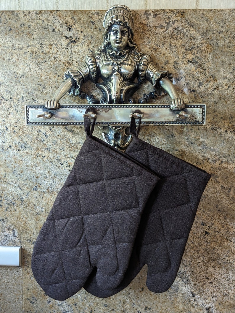 K&ouml;nnte beinhalten: Zwei braune, gesteppte Ofenhandschuhe, die an einem silbernen Metallgestell h&auml;ngen, auf dem sich eine dekorative Figur einer Frau in einem Kleid befindet.