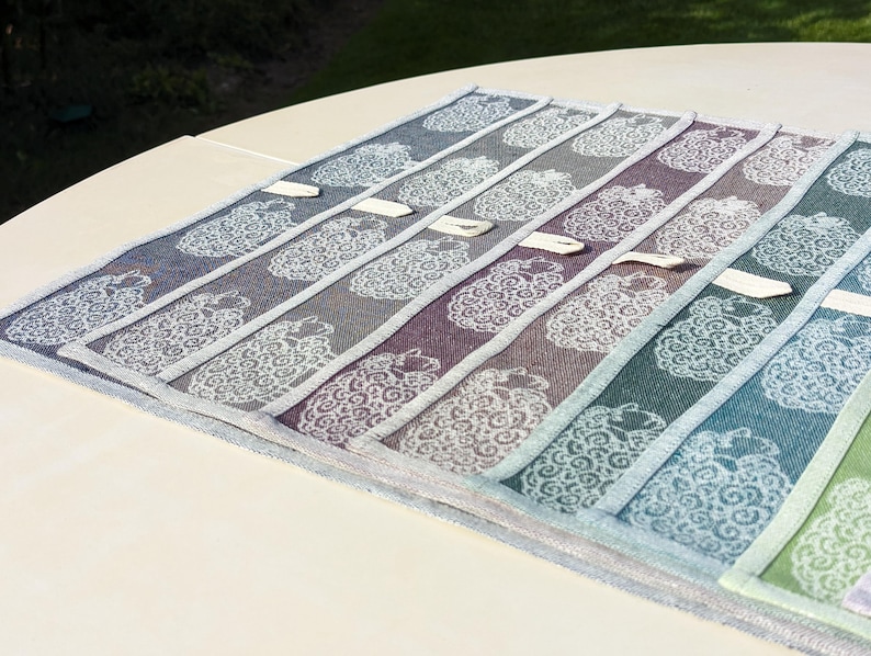 May include: A set of ten kitchen towels featuring a sheep design in shades of blue, green, purple, and grey. The towels are made from linen and have a fringed edge.