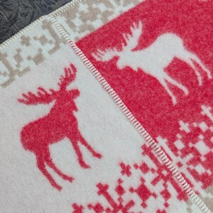 May include: A white and red woven blanket with a moose design. The blanket has a beige border and a white snowflake pattern.