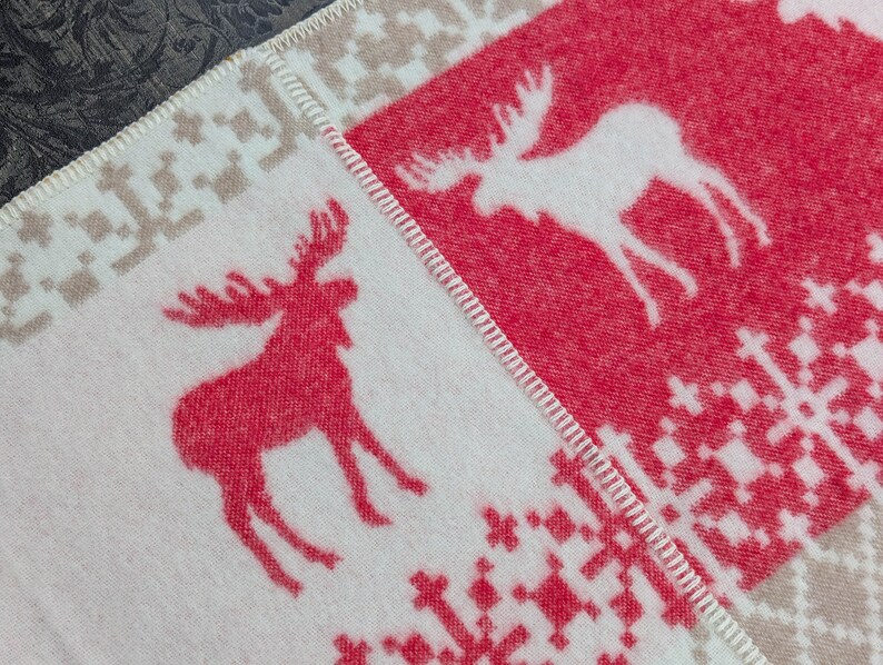 May include: A white and red woven blanket with a moose design. The blanket has a beige border and a white snowflake pattern.