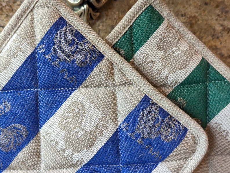 Puede incluir: Dos agarraderas acolchadas, una azul y blanca y otra verde y blanca, con un patr&oacute;n de p&aacute;jaros estilizados.