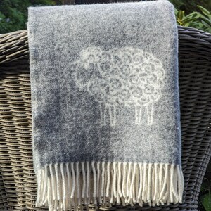 May include: A gray wool throw blanket with a white sheep design and fringe. The blanket is draped over a wicker chair.