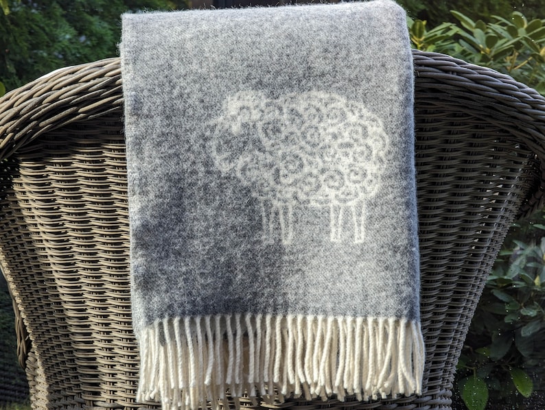 May include: A gray wool throw blanket with a white sheep design and fringe. The blanket is draped over a wicker chair.