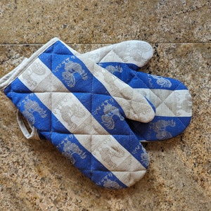 Puede incluir: Un par de guantes de horno a rayas azules y blancas con un dise&ntilde;o acolchado. Los guantes tienen un patr&oacute;n de gallos estilizados.