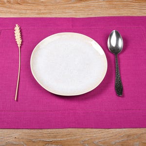 May include: A pink linen placemat with a white plate, a silver spoon, and a wheat stalk.
