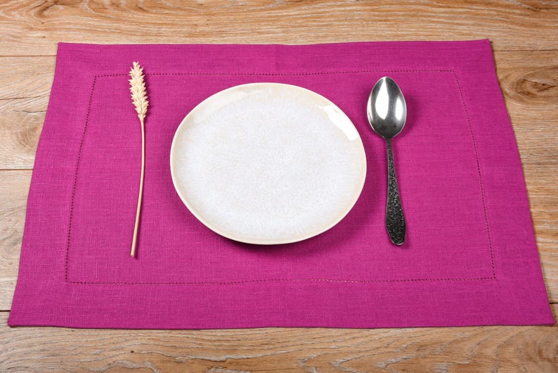 May include: A pink linen placemat with a white plate, a silver spoon, and a wheat stalk.