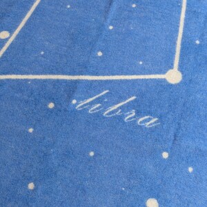 May include: A blue fabric background with white stars and a white outline of the constellation Libra. The word "Libra" is written in white cursive lettering.