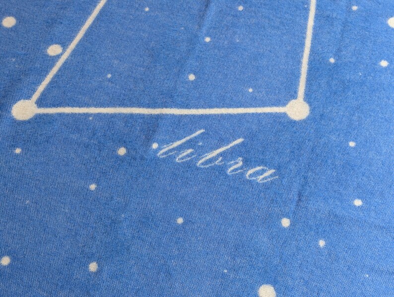 May include: A blue fabric background with white stars and a white outline of the constellation Libra. The word "Libra" is written in white cursive lettering.