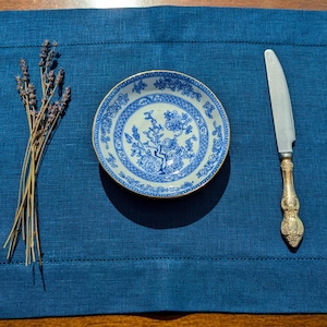 May include: A blue and white porcelain plate with a floral design sits on a blue linen placemat with a silver knife and a bunch of lavender.
