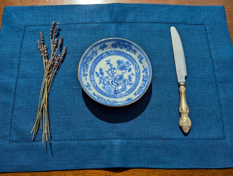May include: A blue and white porcelain plate with a floral design sits on a blue linen placemat with a silver knife and a bunch of lavender.