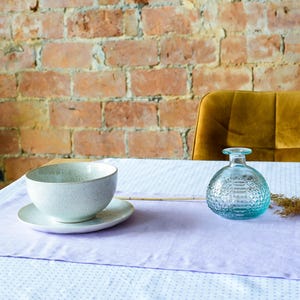May include: A light purple table runner with a white background and small white dots. A white bowl with a gray speckled design sits on the runner, along with a clear glass bottle with a textured design. A dried flower stalk is placed on the runner next to the bottle.