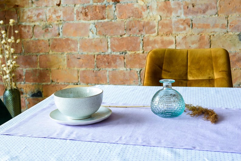 May include: A light purple table runner with a white background and small white dots. A white bowl with a gray speckled design sits on the runner, along with a clear glass bottle with a textured design. A dried flower stalk is placed on the runner next to the bottle.