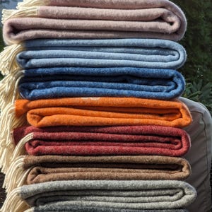 May include: A stack of eight folded blankets in various colors, including pink, blue, orange, red, brown, gray, and dark gray. The blankets are folded neatly and have a white fringe along the edges.