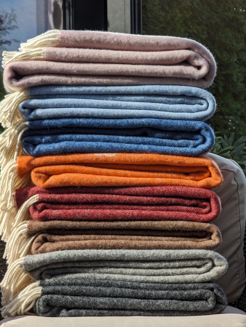 May include: A stack of eight folded blankets in various colors, including pink, blue, orange, red, brown, gray, and dark gray. The blankets are folded neatly and have a white fringe along the edges.