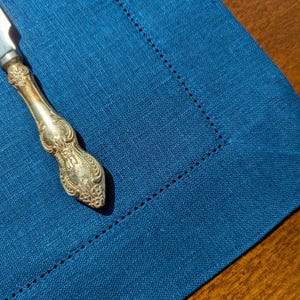 May include: A silver butter knife with an ornate handle rests on a dark blue linen placemat.