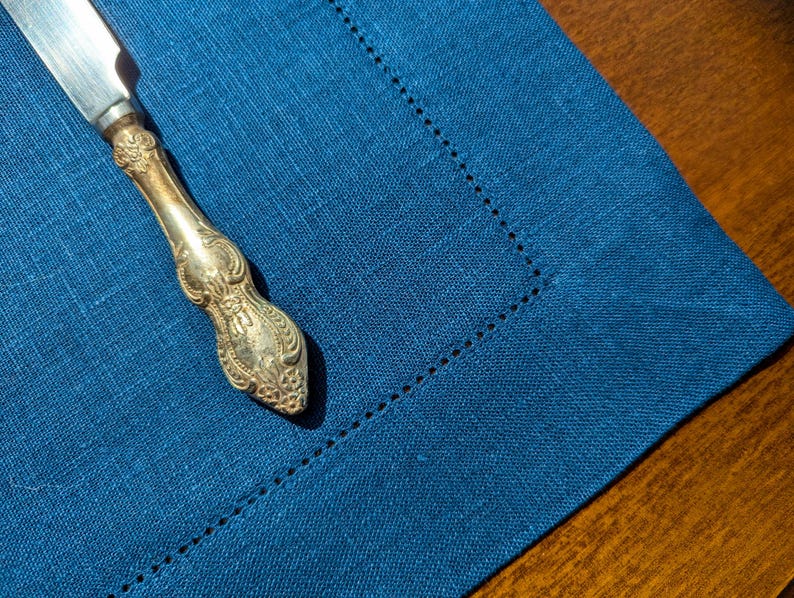 May include: A silver butter knife with an ornate handle rests on a dark blue linen placemat.