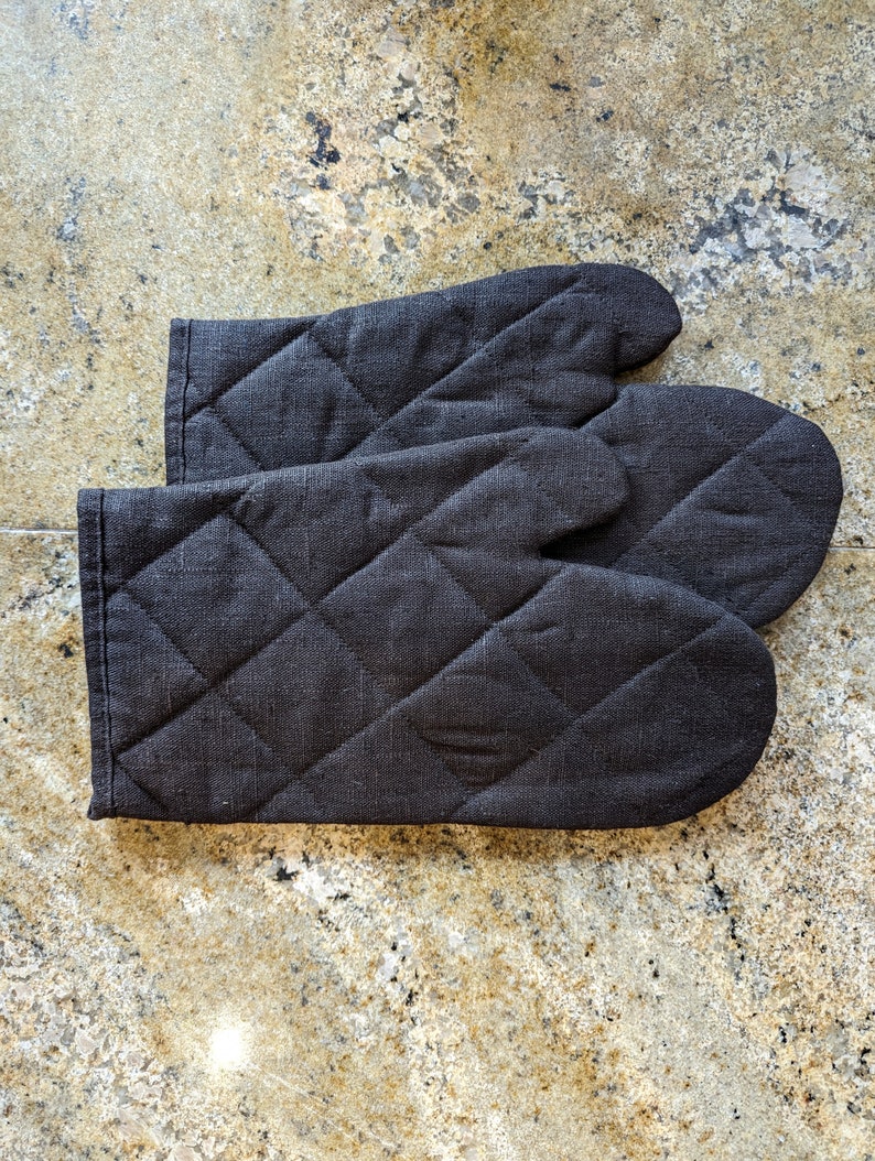 K&ouml;nnte beinhalten: Zwei schwarze, gesteppte Ofenhandschuhe, die auf einer hellbraunen, gesprenkelten Arbeitsplatte zusammengelegt sind.