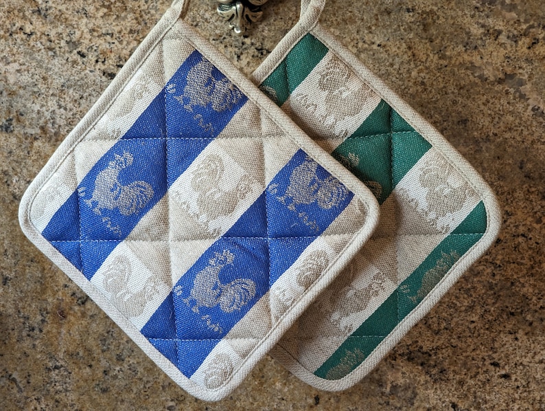 Puede incluir: Dos agarraderas acolchadas con un dise&ntilde;o de gallo. Una agarradera es azul y blanca, y la otra es verde y blanca.
