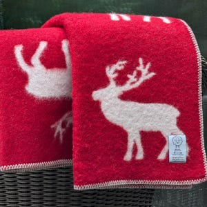 May include: Red wool throw blanket with a white reindeer design. The blanket has a white border and a small white tag with the text "Made in Scotland" on it.