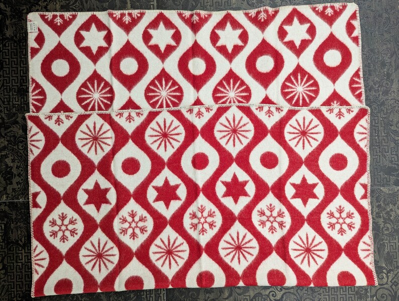 Puede incluir: Dos mantas estampadas en rojo y blanco. El dise&ntilde;o presenta un patr&oacute;n repetitivo de gotas, estrellas, c&iacute;rculos y copos de nieve rojos sobre un fondo blanco. Los bordes est&aacute;n acabados con un borde cosido blanco. Las mantas est&aacute;n dobladas y apiladas.