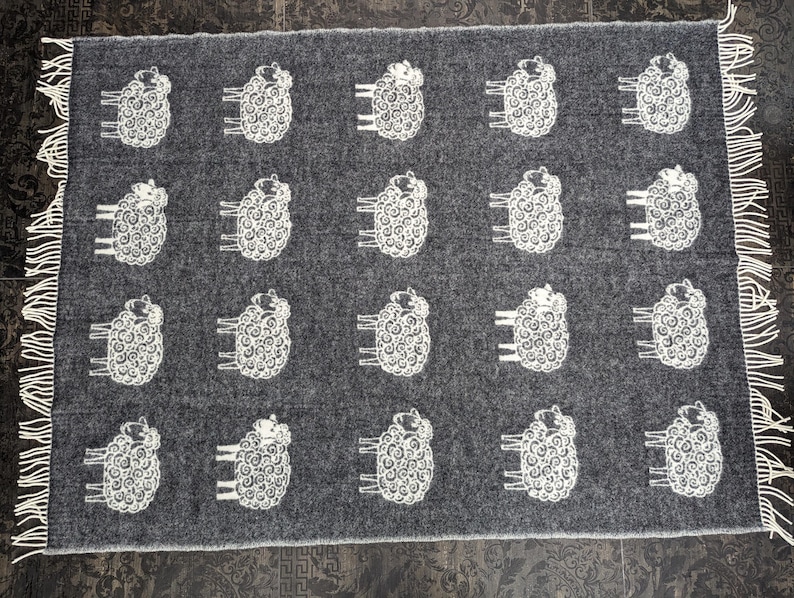 May include: A gray wool throw blanket with a white sheep design. The blanket has fringe on all sides.