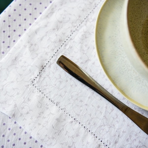 May include: A dining table setting with a white and grey floral patterned napkin with a stitched border, a plate, and a bowl. A silver knife rests on the napkin. The tablecloth has a white background with purple polka dots.