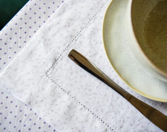 May include: A table setting featuring a white and gray floral patterned napkin with a stitched border, a plate, and a bowl. A silver knife rests on the napkin. The tablecloth has a white background with purple polka dots.