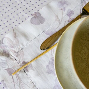 May include: A close-up of a light brown ceramic bowl with a speckled glaze. The bowl is sitting on a white tablecloth with a floral pattern. A silver fork and a wooden skewer are also visible in the image.