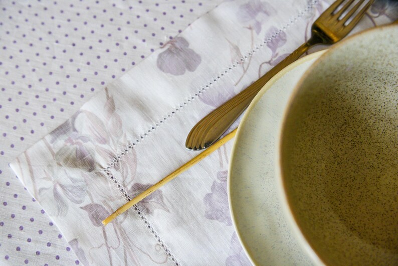 May include: A close-up of a light brown ceramic bowl with a speckled glaze. The bowl is sitting on a white tablecloth with a floral pattern. A silver fork and a wooden skewer are also visible in the image.