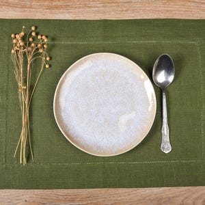 May include: A green linen placemat with a white ceramic plate and a silver spoon on top.