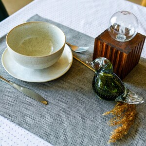 May include: A gray ceramic bowl with a white saucer, a silver knife, and a glass bird figurine on a gray placemat. A wooden base with a glass light bulb sits on the table.