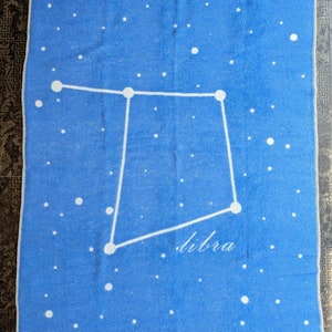May include: A blue blanket with a white constellation of Libra printed on it. The blanket is decorated with white dots to represent stars.