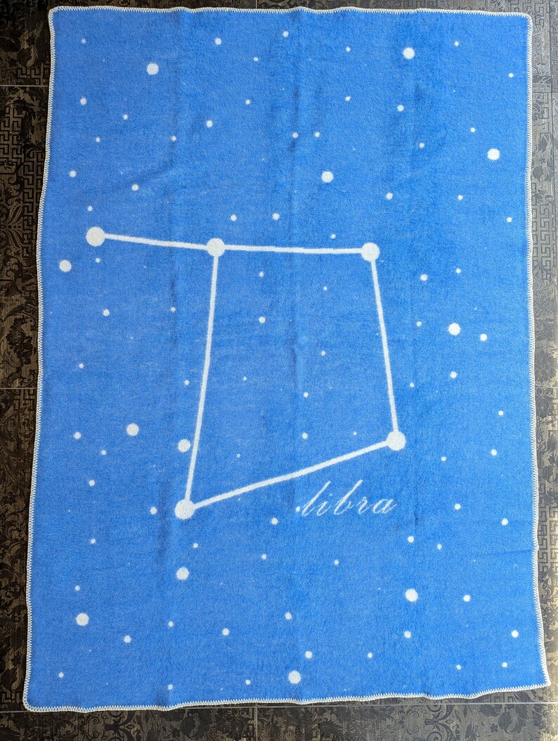 May include: A blue blanket with a white constellation of Libra printed on it. The blanket is decorated with white dots to represent stars.