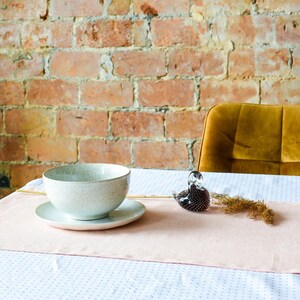 May include: A white ceramic bowl with a speckled glaze sits on a white saucer on a pink table runner. The table runner is on a white tablecloth with a subtle pattern. A glass bird figurine is on the table to the right of the bowl.