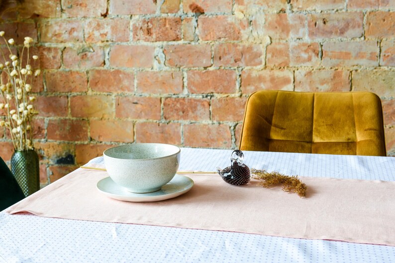May include: A white ceramic bowl with a speckled glaze sits on a white saucer on a pink table runner. The table runner is on a white tablecloth with a subtle pattern. A glass bird figurine is on the table to the right of the bowl.