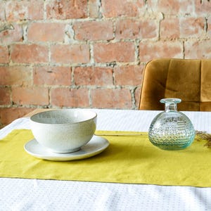 May include: A yellow table runner with a white bowl and a clear glass bottle with a textured surface. The table runner is on a white tablecloth with a small pattern.