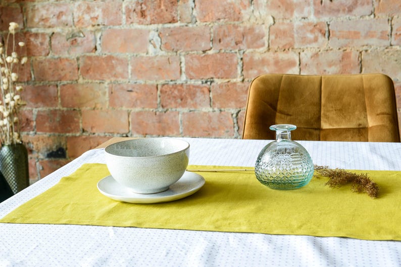 May include: A yellow table runner with a white bowl and a clear glass bottle with a textured surface. The table runner is on a white tablecloth with a small pattern.
