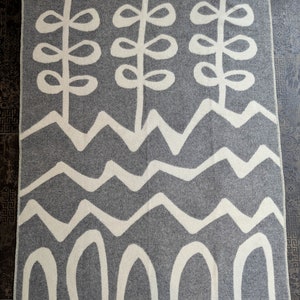 May include: A gray and white wool throw blanket with a geometric pattern of stylized plants, waves, and arches.