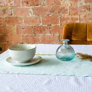 May include: A light blue table runner with a white background and a small pattern of white dots. A white ceramic bowl, a clear glass bottle with a textured surface, and a dried flower stem are on the table runner.