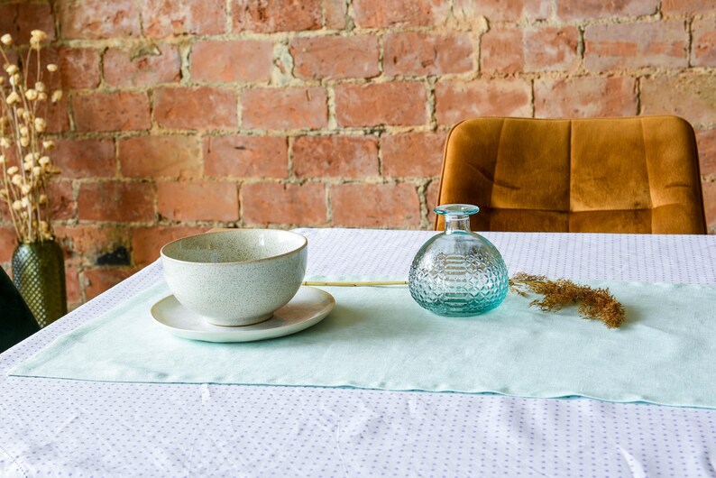May include: A light blue table runner with a white background and a small pattern of white dots. A white ceramic bowl, a clear glass bottle with a textured surface, and a dried flower stem are on the table runner.