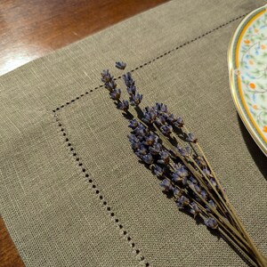 Puede incluir: Un mantel individual de lino beige con un borde decorativo cosido. Una rama de lavanda seca se coloca sobre el mantel, y un plato de porcelana blanca con un dise&ntilde;o floral est&aacute; en el fondo.